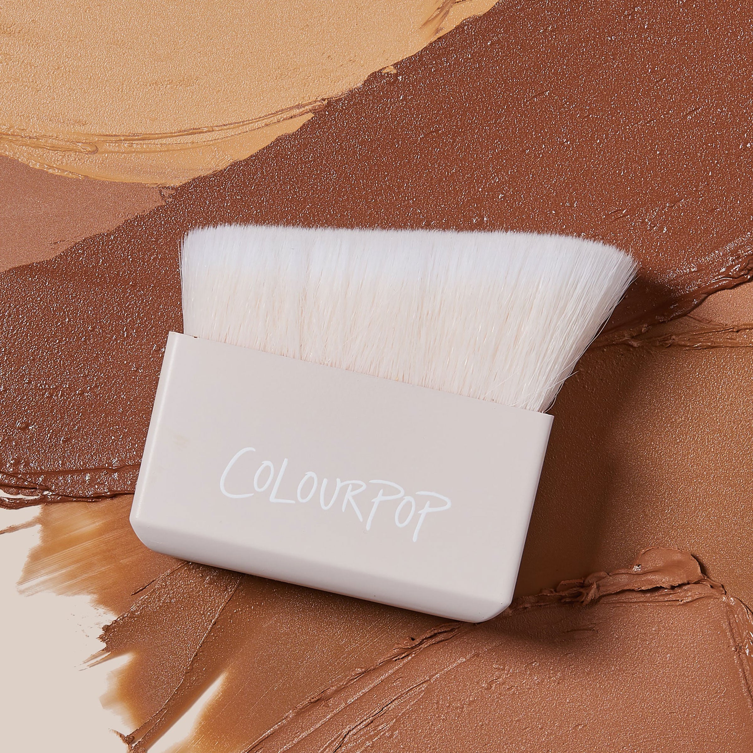 ColourPop Makeup Brush Angled Sculpting Brush resting on swatches of tan to deep brown cream contour; dense white angled bristles extend from a beige block handle stamped ColourPop, ideal for precise cheekbone sculpting.