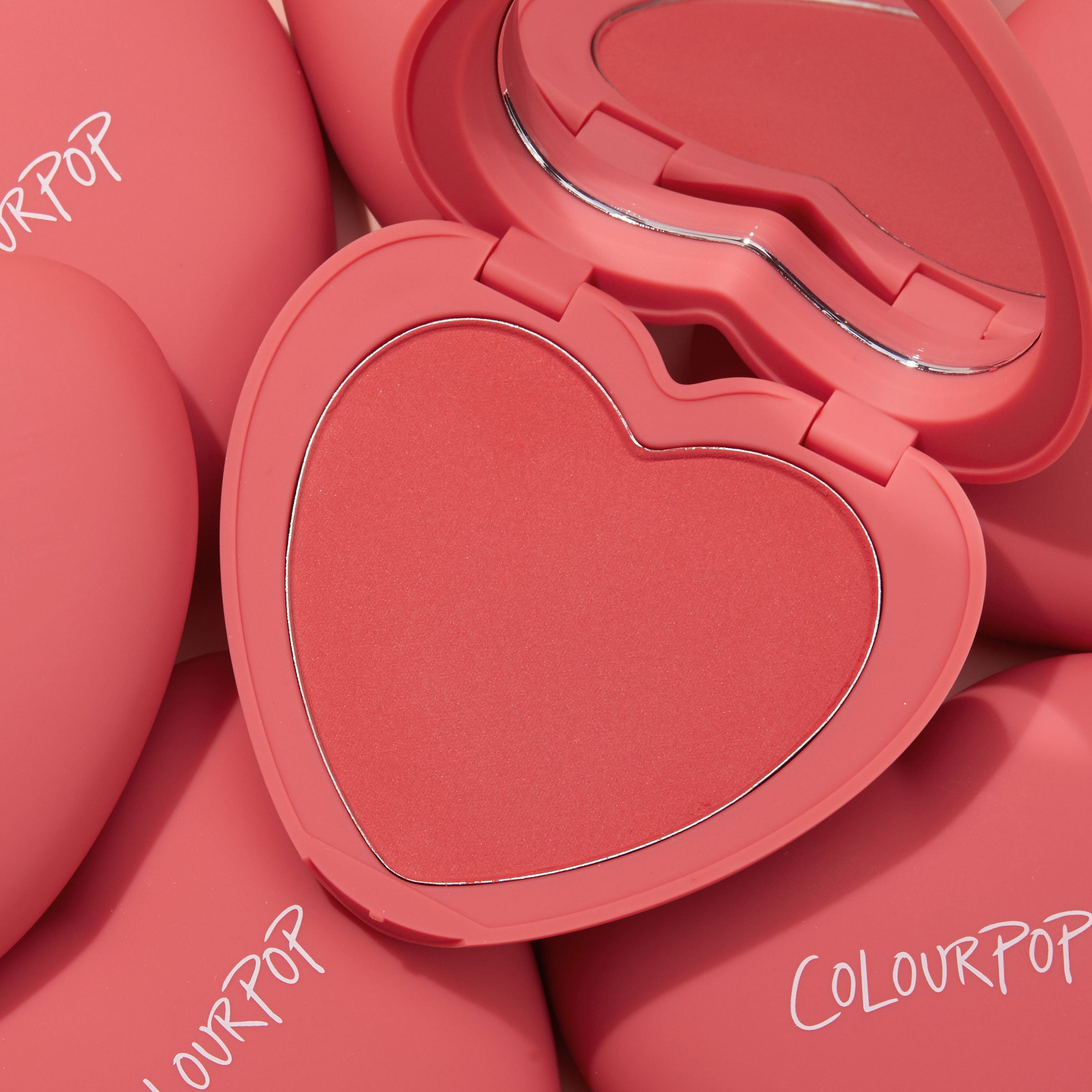 4ever Yours ColourPop Pressed Powder Blush compact opens to reveal a heart-shaped coral pan and mirror, set atop several matte pink cases with ColourPop logos, highlighting the romantic, travel-friendly pressed powder blush.