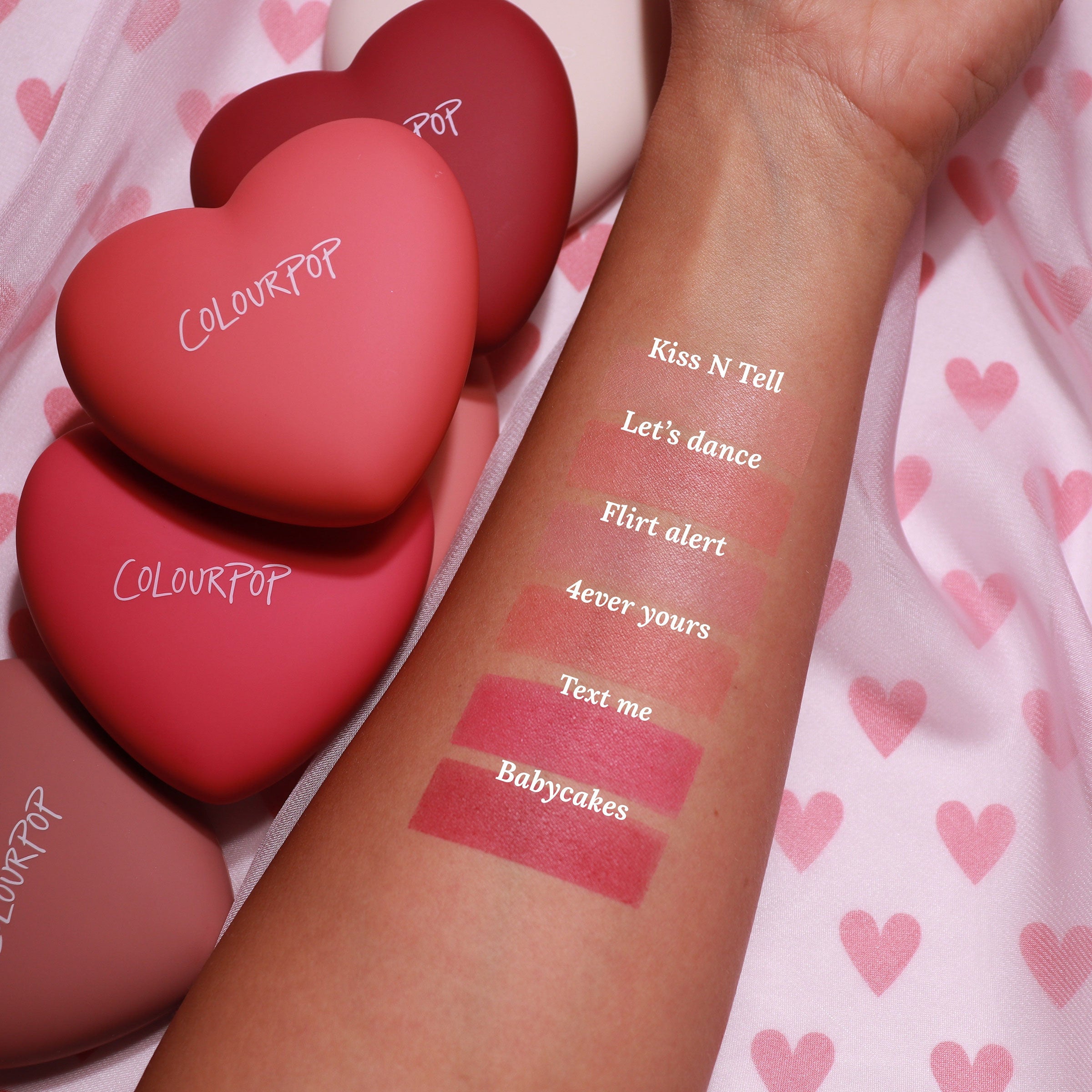 Forearm displays six swatches of ColourPop Pressed Powder Blush, highlighting the 4ever Yours shade among Kiss N Tell, Let’s Dance, Flirt Alert, Text Me, and Babycakes, next to heart-shaped ColourPop compacts on pink heart-print fabric.