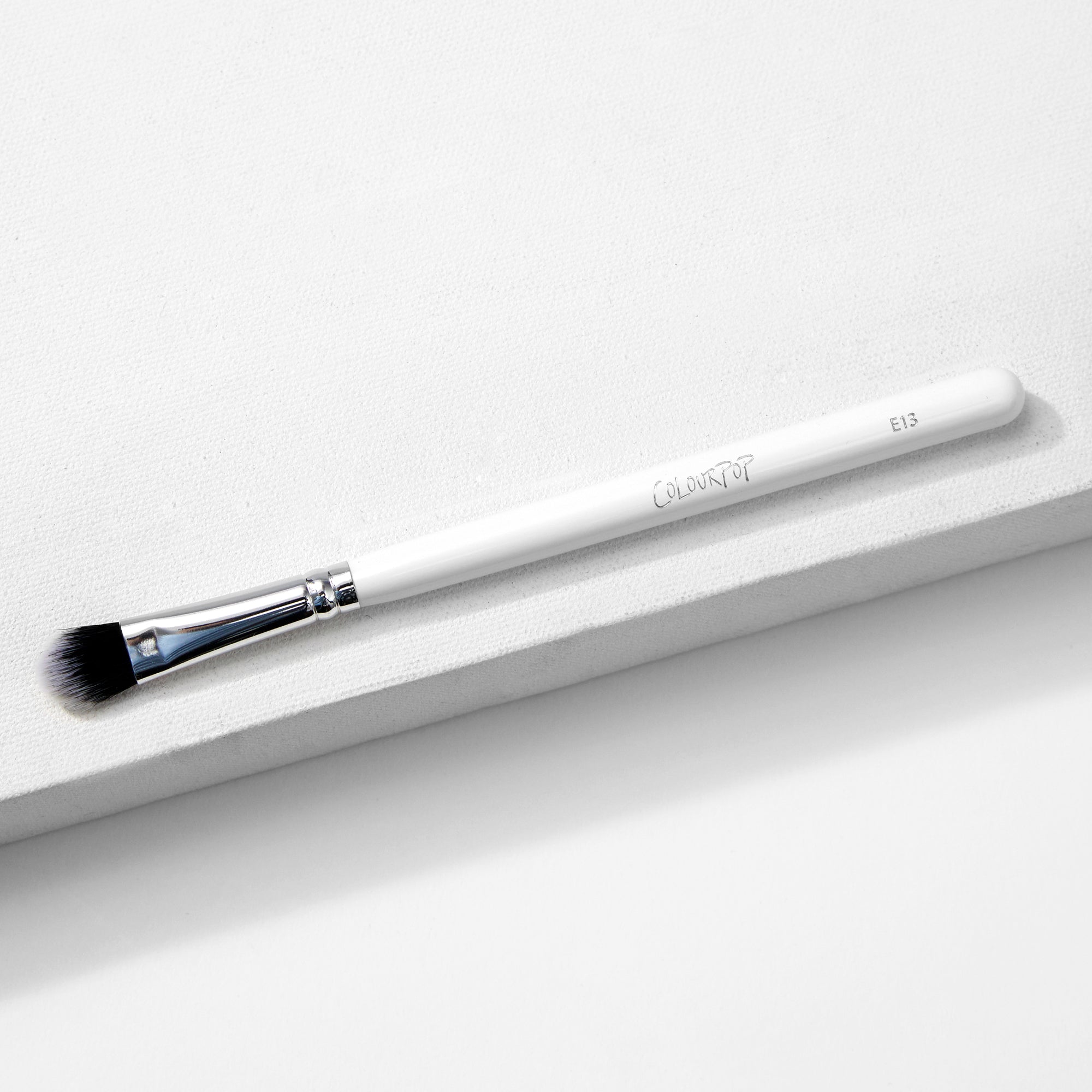 Large Shader Brush makeup brush by ColourPop lies diagonally on a white textured surface, showcasing a white handle with logo and E13, a shiny silver ferrule, and flat dense black-to-white bristles designed for packing and shading eyeshadow.